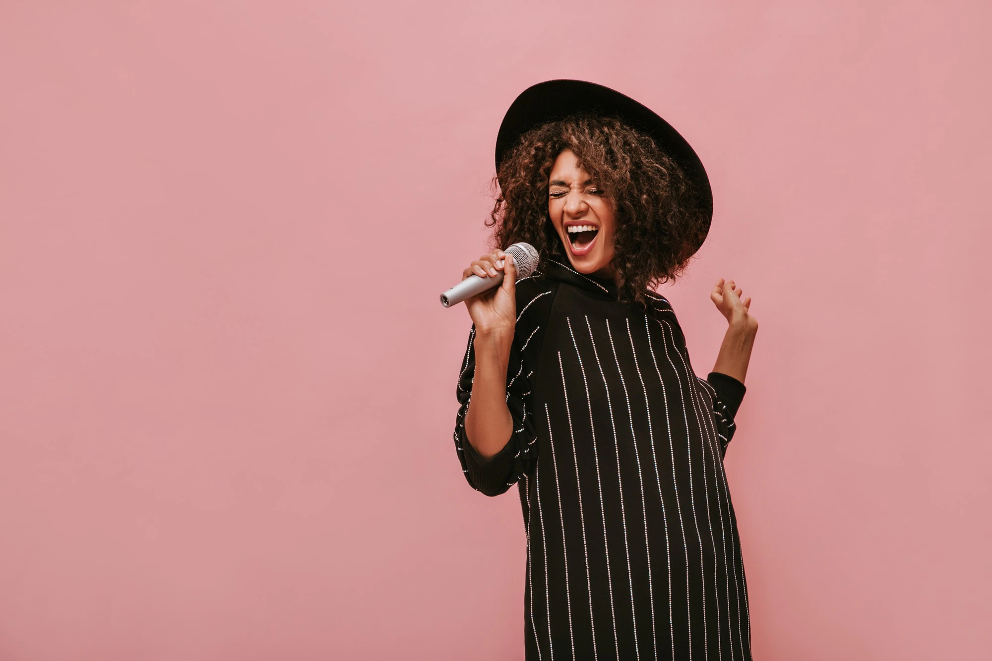 emotional woman with curly brunette hairstyle in stylish hat and striped black dress holding microphone and singing on pink wall 197531 23468