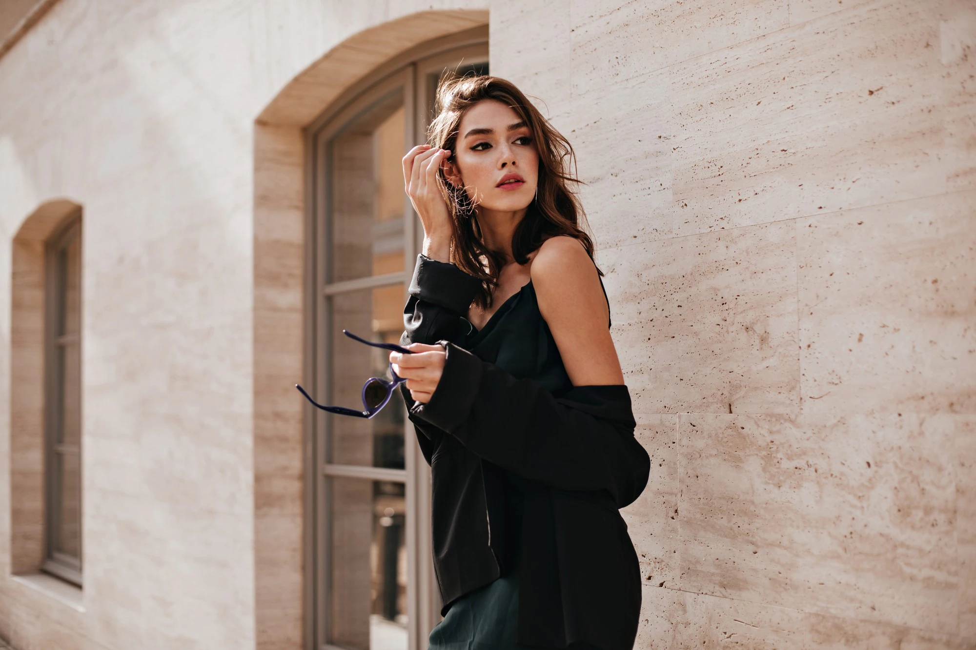 cute young girl with dark wavy hairstyle bright makeup silk dress black jacket holding sunglasses hands looking away against beige building wall 197531 24462