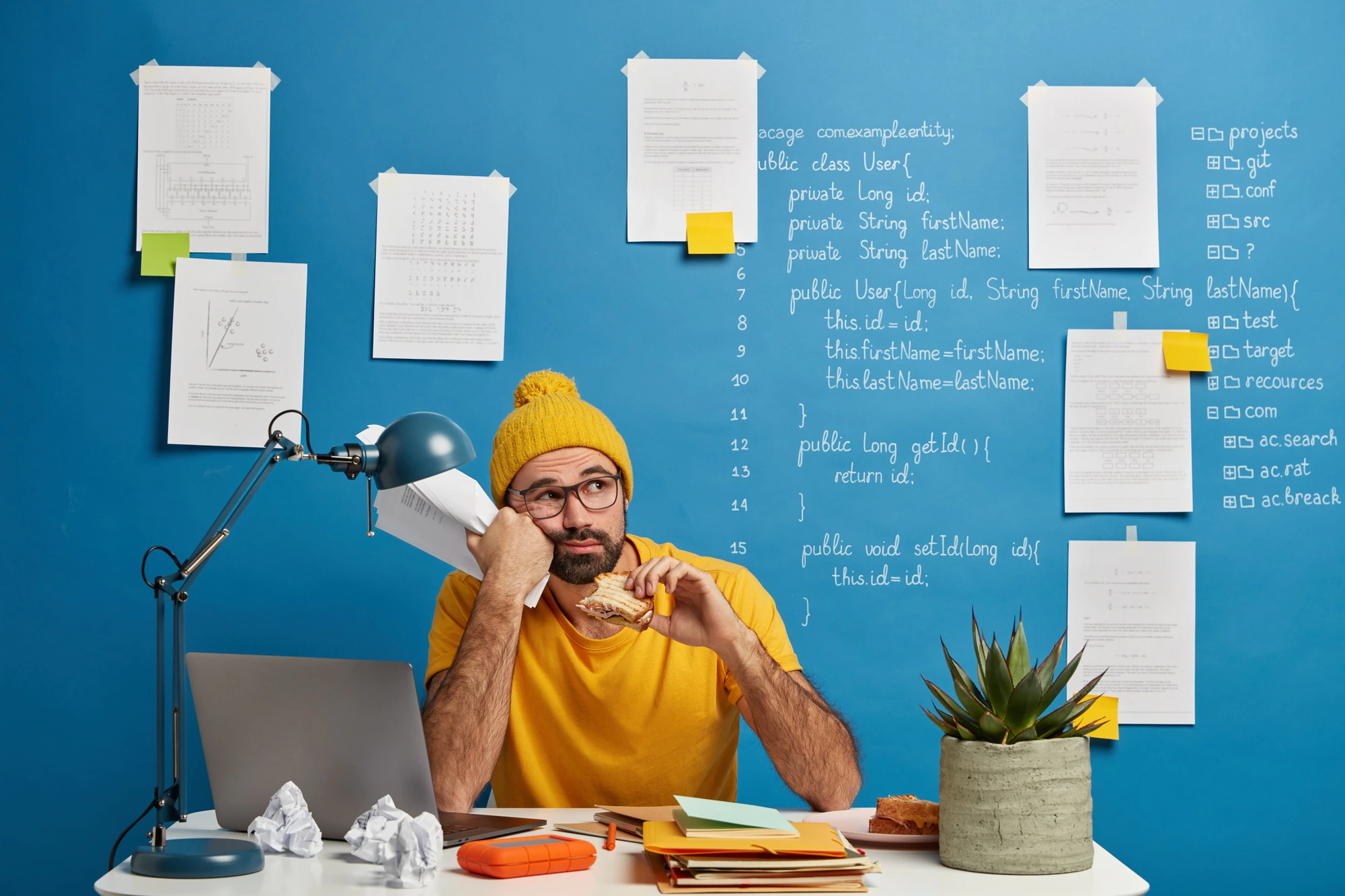 thoughtful male programmer software developer ponders program code looks away eats burger holds papers wears yellow clothes spends time making project 273609 34356